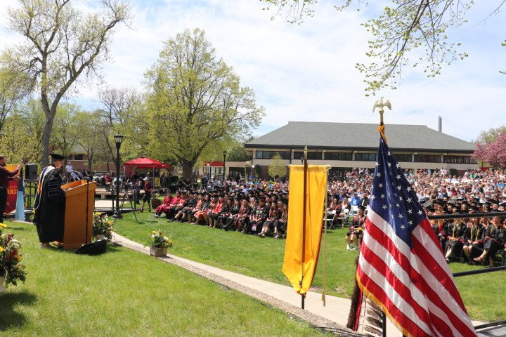 A graduation ceremony taking place outdoors on a sunny day. A speaker stands at a podium addressing a crowd of graduates seated in caps and gowns, while the audience watches from the lawn. An American flag and a yellow banner are visible in the foreground.